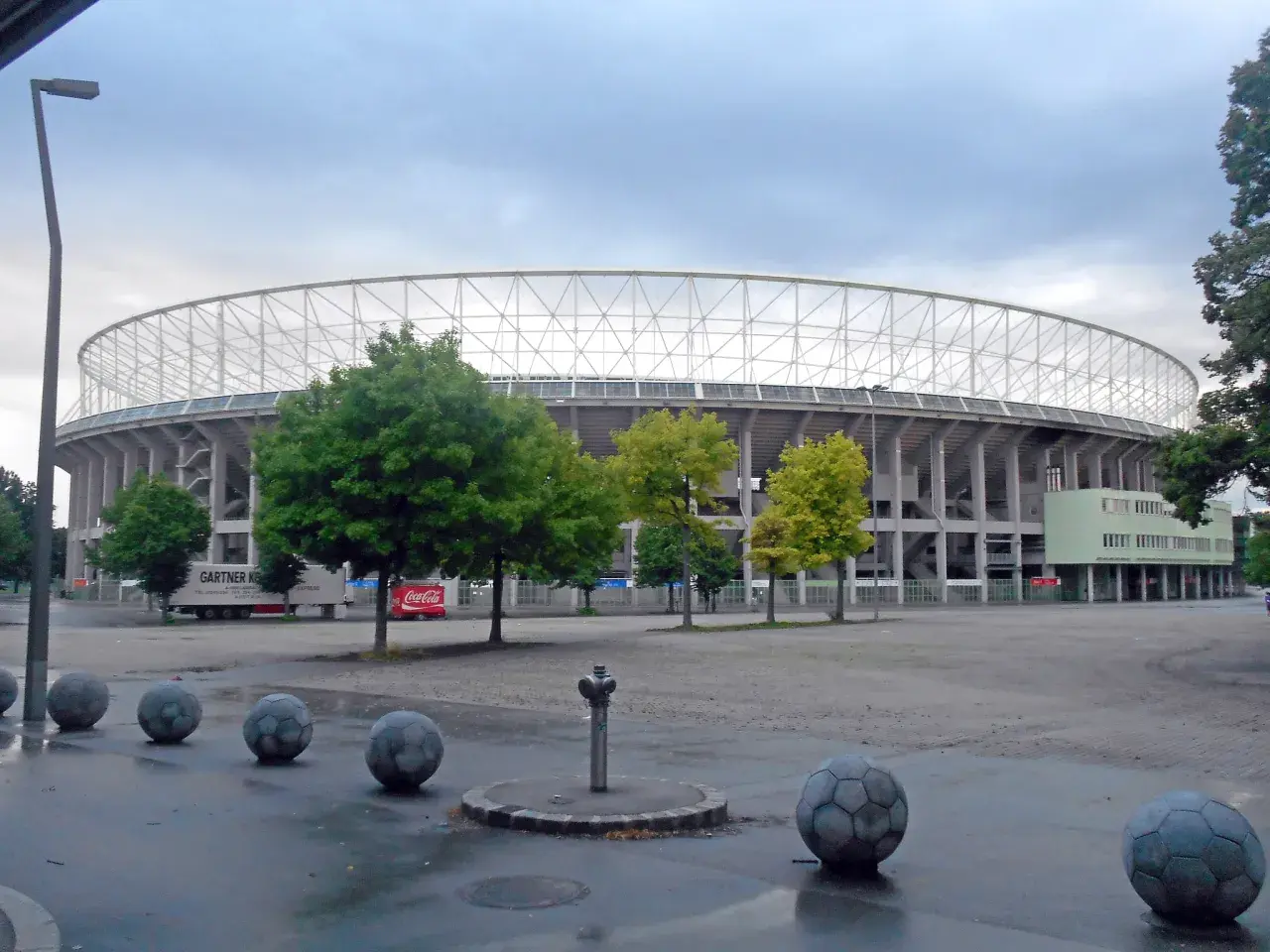 Ernst Happel Stadion z charakterystyczną białą kratownicą dachu, otoczony zielenią i betonowymi kulami przypominającymi piłki nożne.