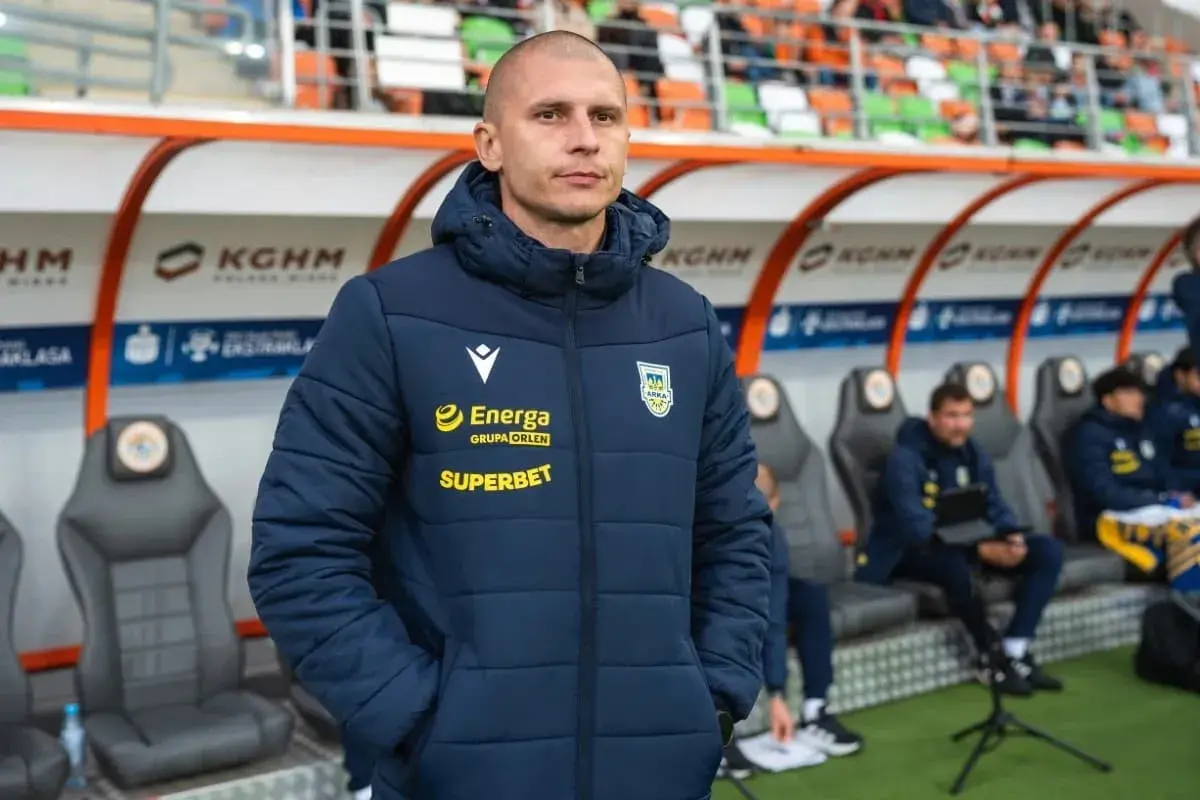 Trener Arki Gdynia, w kurtce z logo Energa i Superbet, stoi na tle ławki rezerwowych.