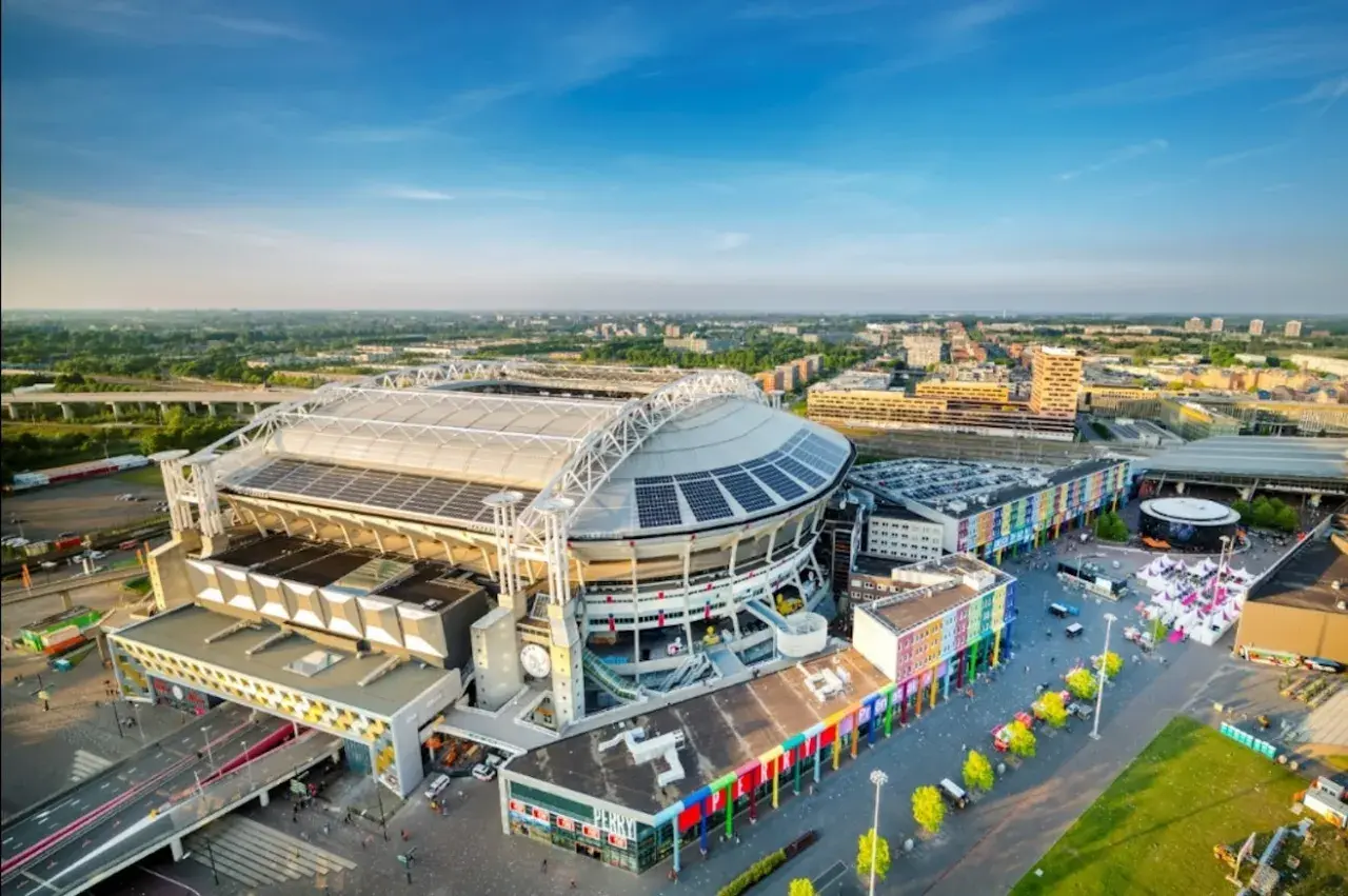 Johan Cruyff Arena, imponujący stadion z panelami słonecznymi na dachu, otoczony kolorowymi budynkami i zielenią.
