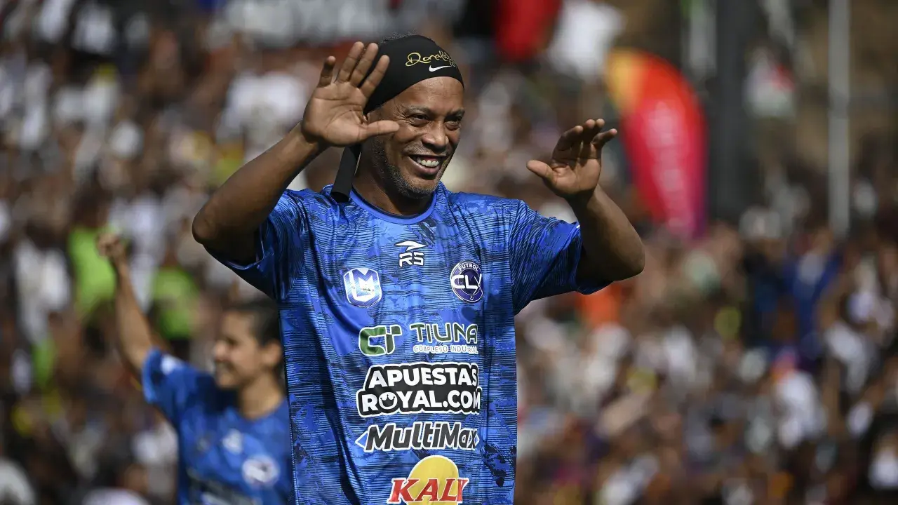 Uśmiechnięty Ronaldinho w niebieskiej koszulce z logo "FUTBOL CL" macha do kibiców. Widać, gdzie gra Ronaldinho.