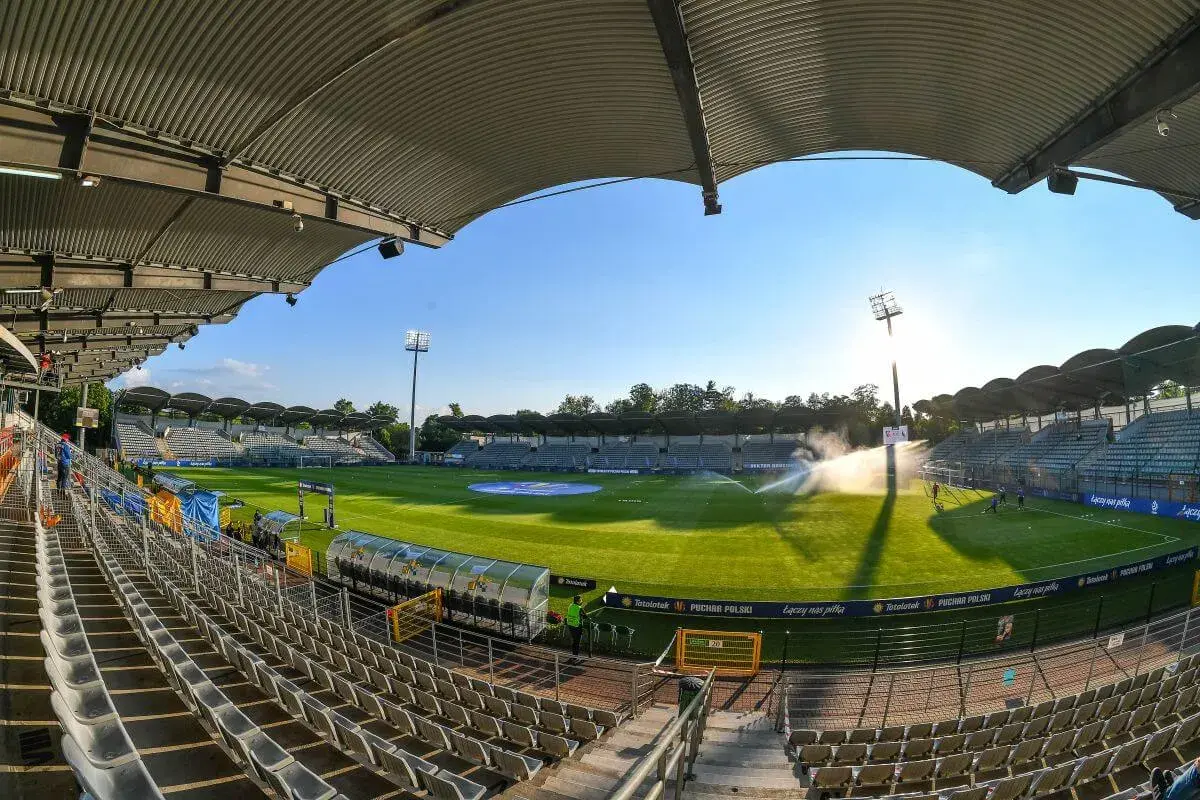 Pusta trybuna stadionu Miedzi Legnica, zielona murawa i zraszacze tworzą spokojny obraz przed meczem.