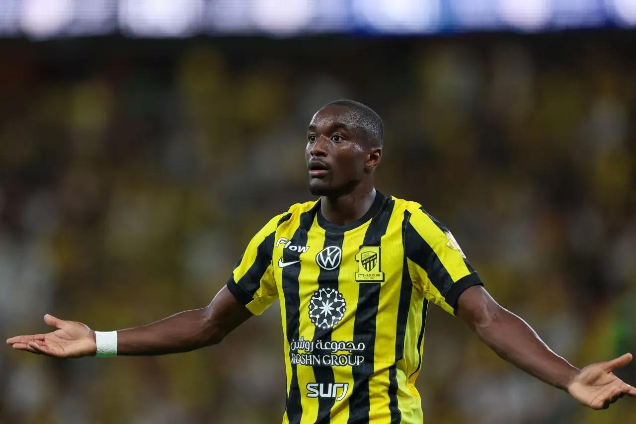 Piłkarz Moussa Diaby w żółto-czarnej koszulce z logo Iттihad Club, z otwartymi dłońmi, gotowy do gry.