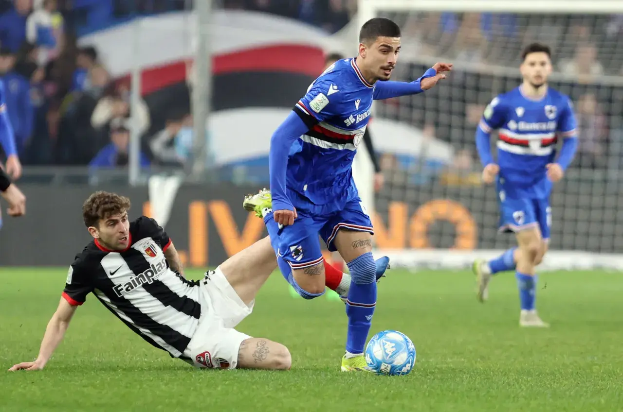 Piłkarze w akcji podczas rozgrywek Serie B. Jeden z zawodników w niebieskim stroju próbuje odebrać piłkę leżącemu na trawie przeciwnikowi w czarno-białej koszulce.