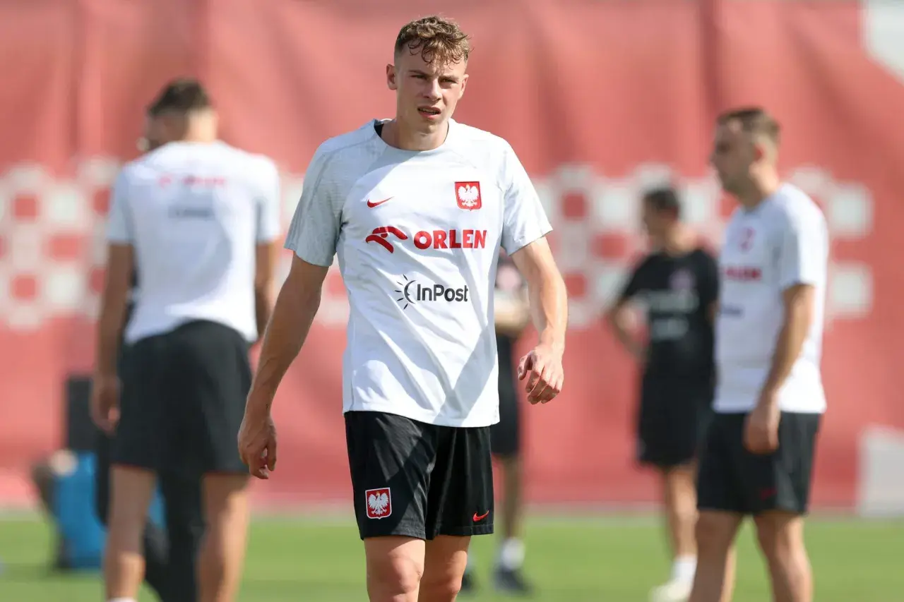 Szymon Żurkowski w białej koszulce reprezentacji Polski z logo Orlen i InPost, na tle czerwonej bandy.