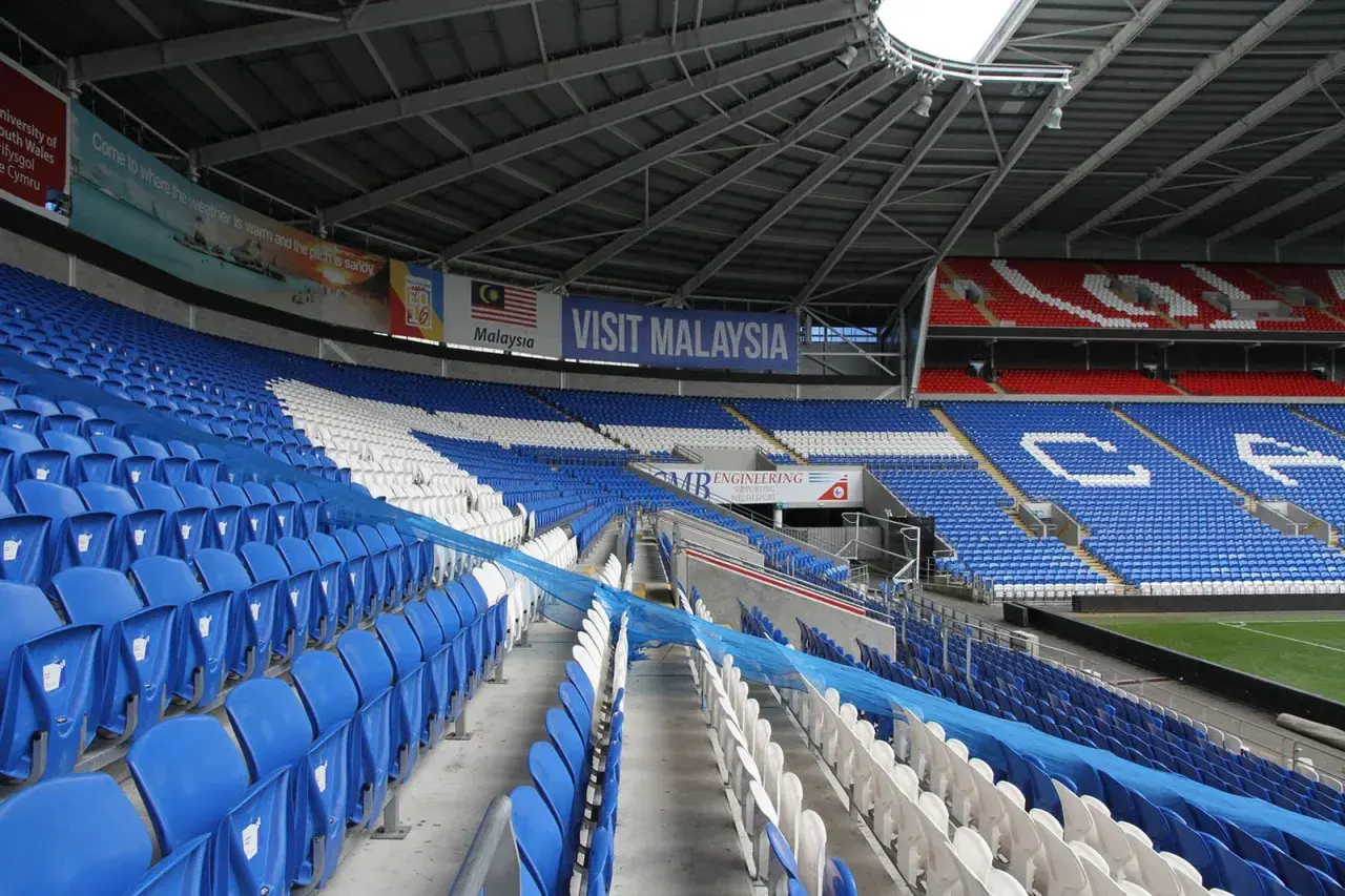 Puste trybuny Cardiff City Stadium, z niebieskimi i białymi siedzeniami, ozdobione banerem "Visit Malaysia".