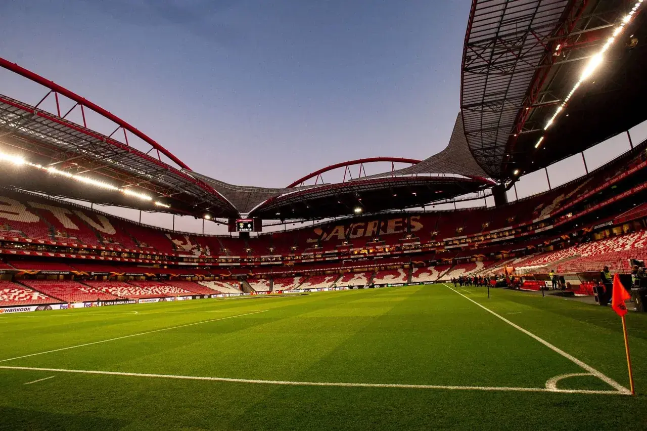 Pusta trybuna stadionu **Estadio da Luz** w Lizbonie, z zieloną murawą i czerwonymi siedzeniami.