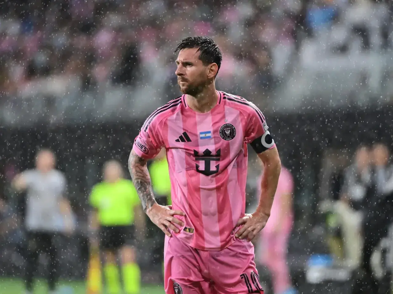 Lionel Messi w różowej koszulce Interu Miami, stojący w deszczu. Wygląda na zamyślonego, być może z powodu niedawnej kontuzji.
