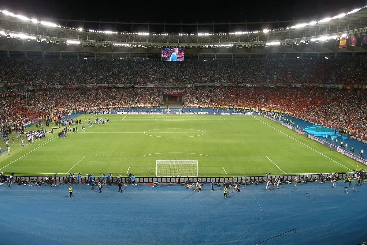 Piłkarze wychodzą na murawę stadionu olimpijskiego w Kijowie. Tłumy kibiców na trybunach, wielki ekran nad boiskiem.