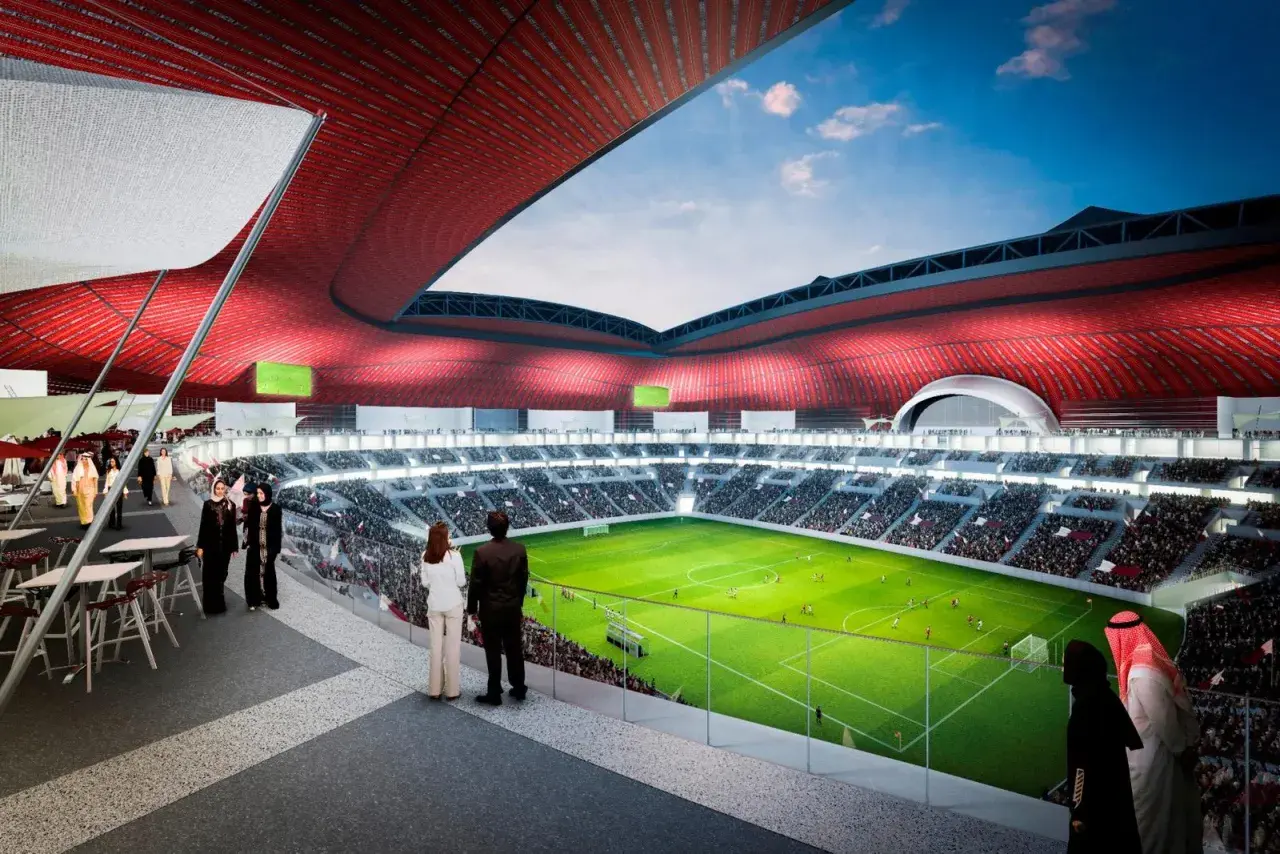 Widok na zieloną murawę stadionu Al Bayt, otoczoną trybunami pełnymi widzów. Czerwony dach tworzy imponującą konstrukcję.