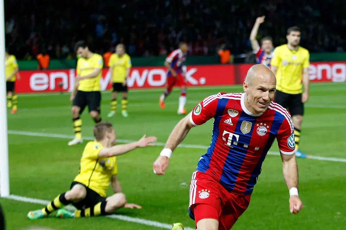 Piłkarz Bayernu w czerwono-niebieskiej koszulce cieszy się z bramki. W tle zawodnicy Borussii Dortmund w żółtych strojach.