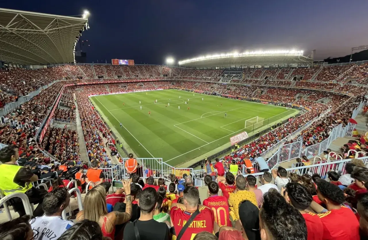 Widok z trybun na pełny stadion **Estadio La Rosaleda** podczas meczu piłki nożnej. Tłum kibiców w czerwonych barwach wypełnia trybuny.