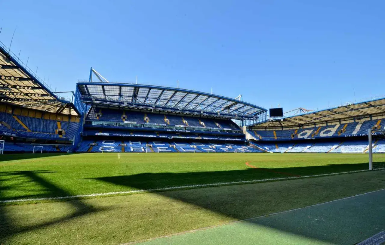 Pusta trybuna nowego stadionu Chelsea, z zieloną murawą i niebieskimi siedzeniami.
