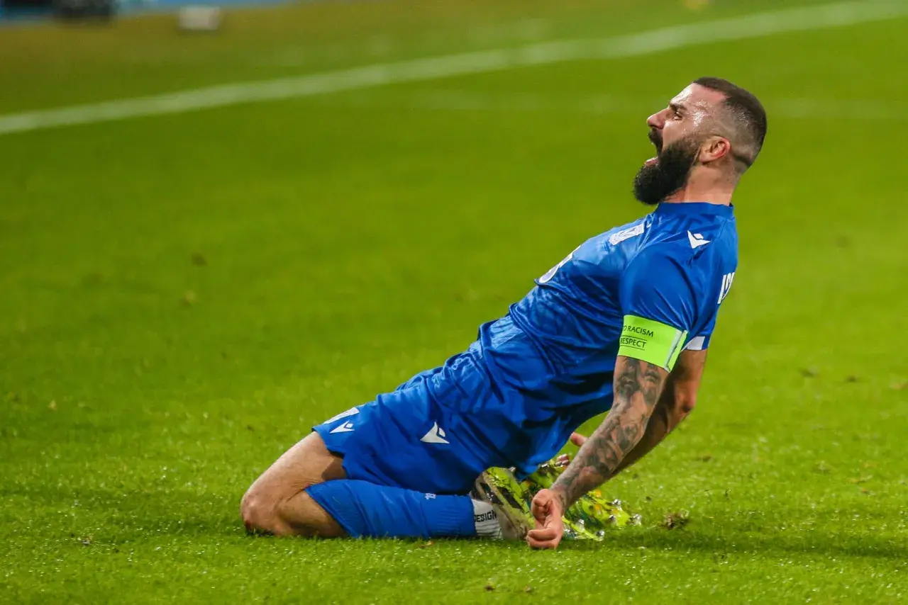 Mikael Ishak świętuje bramkę, klęcząc na zielonej murawie.