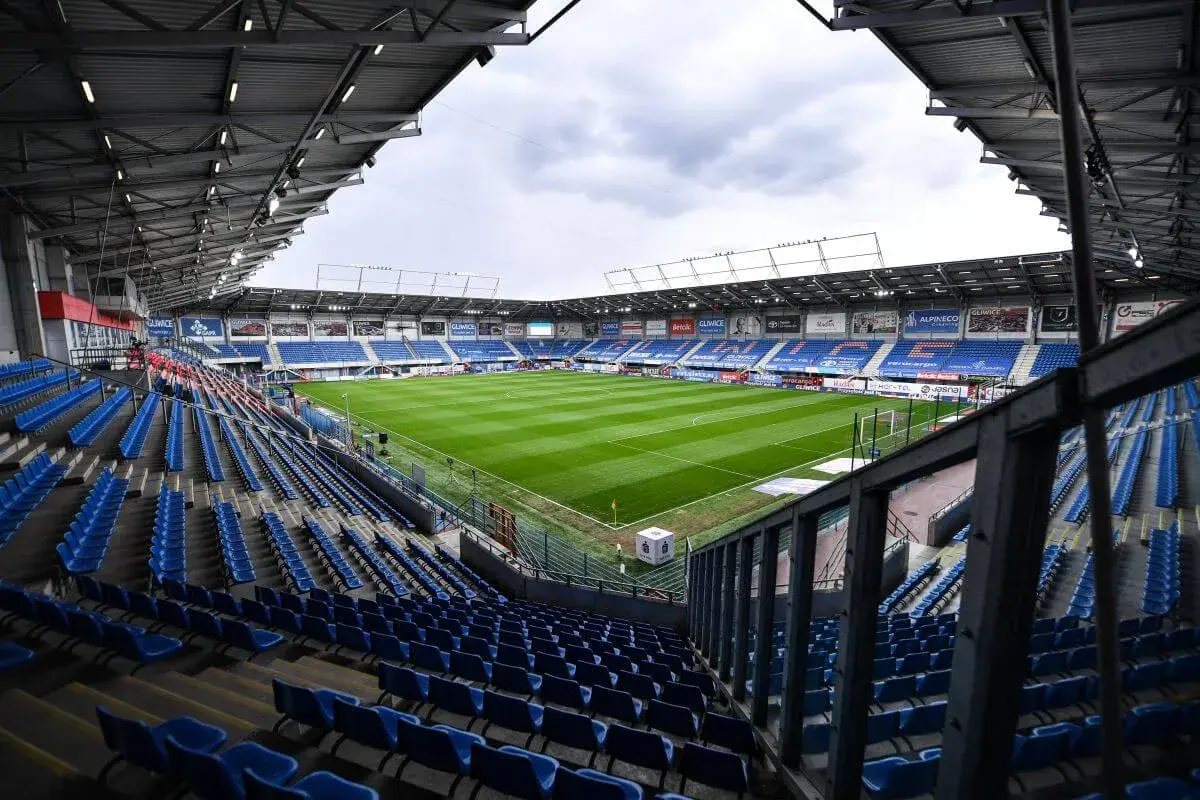 Puste trybuny stadionu Piasta Gliwice czekają na kibiców. Zielona murawa boiska lśni w blasku dnia.