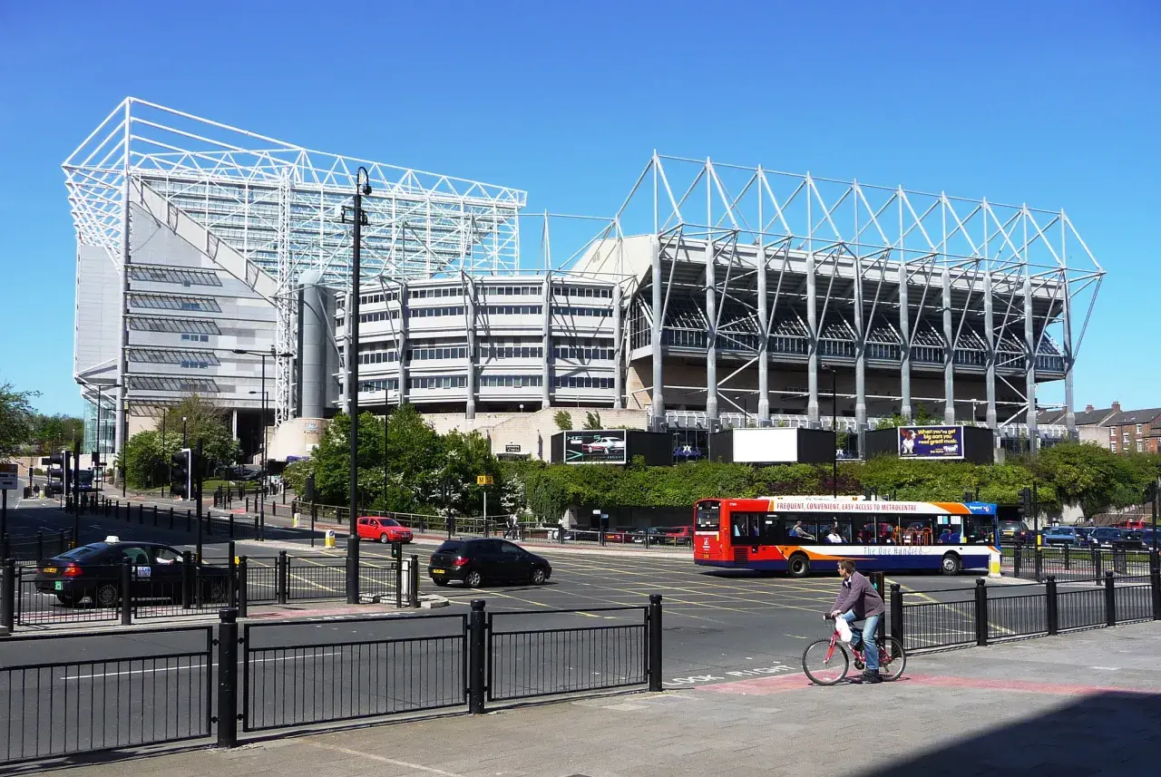 St James' Park w Newcastle, z charakterystyczną białą konstrukcją dachu, otoczony zielenią i ruchliwą ulicą z autobusem i rowerzystą.