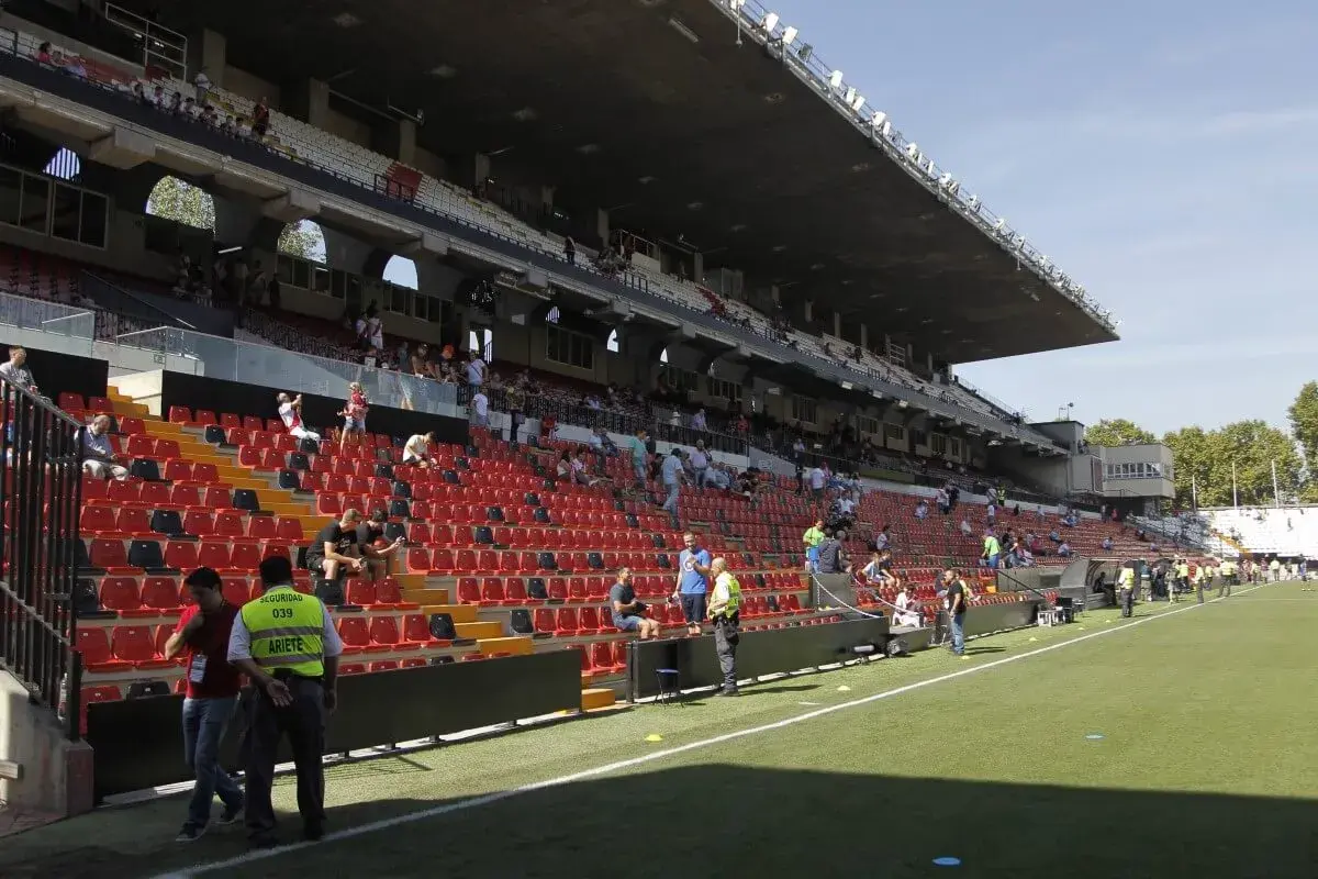 Puste trybuny stadionu Rayo Vallecano czekają na kibiców. Widać zieloną murawę i kilka osób na schodach.