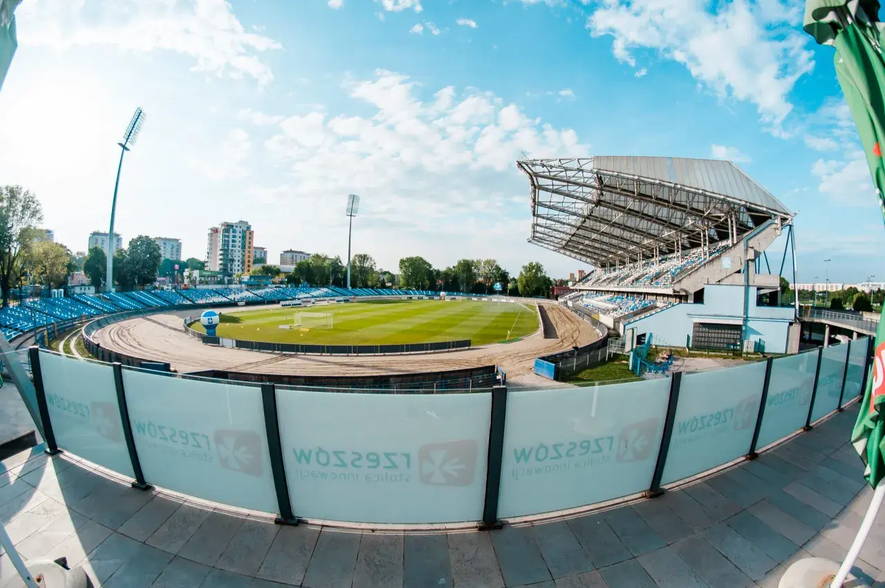 Stadion miejski w Rzeszowie z zieloną murawą, bieżnią żużlową i niebieskimi trybunami. W tle budynki mieszkalne i błękitne niebo z chmurami.