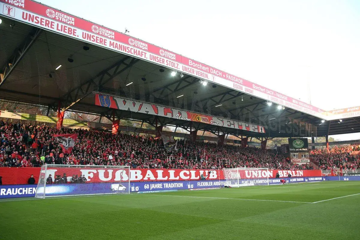 Trybuna na stadionie Union Berlin wypełniona kibicami. Na czerwonym banerze widnieje napis "UNSERE LIEBE. UNSERE MANNSCHAFT. UNSER STOLZ. UNSER VEREIN.".