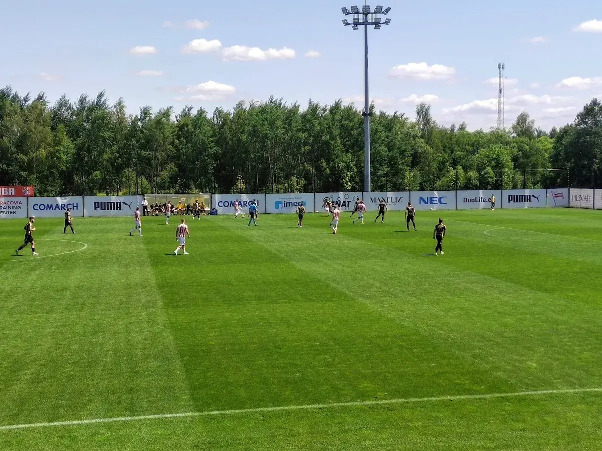 Piłkarze grają na boisku podczas meczu Betclic 3 liga. Słońce świeci, a drzewa tworzą tło.