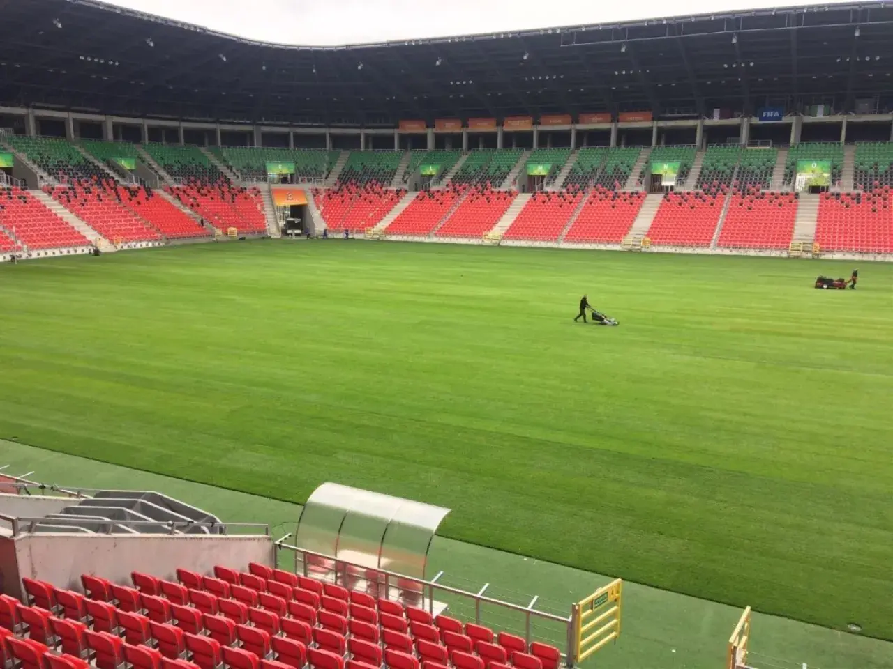 Pracownicy koszą trawę na boisku stadionu GKS Tychy. Puste trybuny w kolorach czerwonym i zielonym czekają na kibiców.