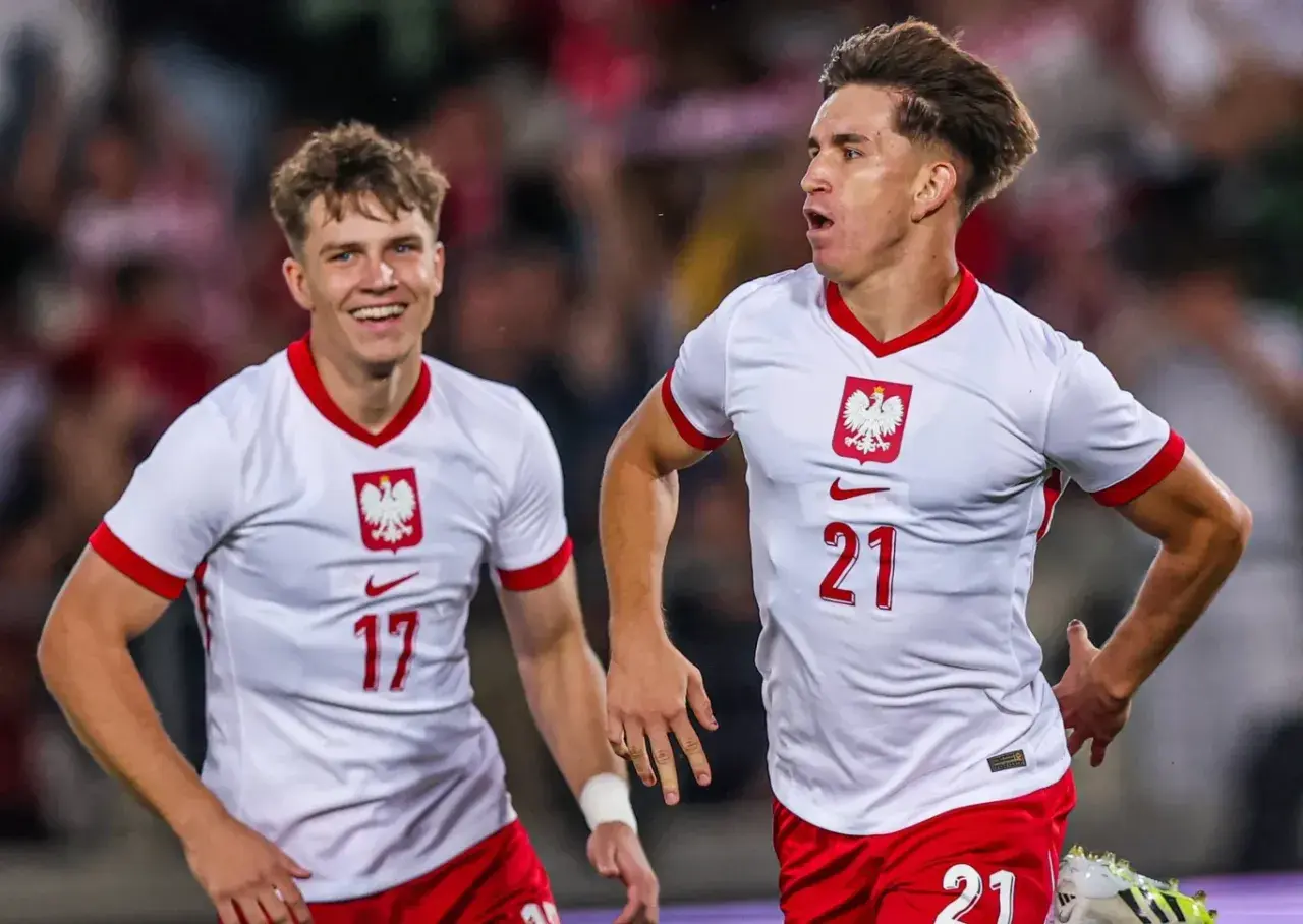 Dwóch młodych zawodników w białych koszulkach z orłem na piersi, z numerami 17 i 21, cieszy się po zdobytej bramce.