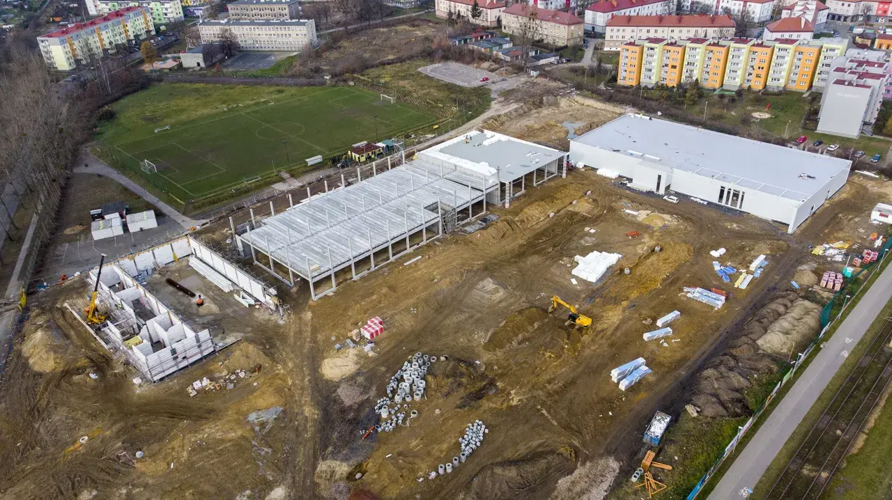 Budowa obiektu sportowego w Radzionkowie. Widać plac budowy z fundamentami, konstrukcją hali, maszynami budowlanymi i pobliski stadion.