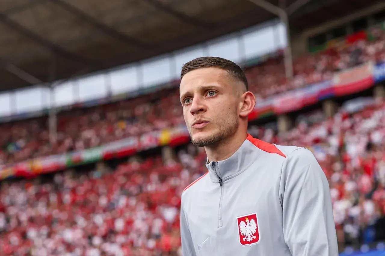 Sebastian Szymański w szarej bluzie z orłem na piersi, na tle trybun wypełnionych kibicami w biało-czerwonych barwach.