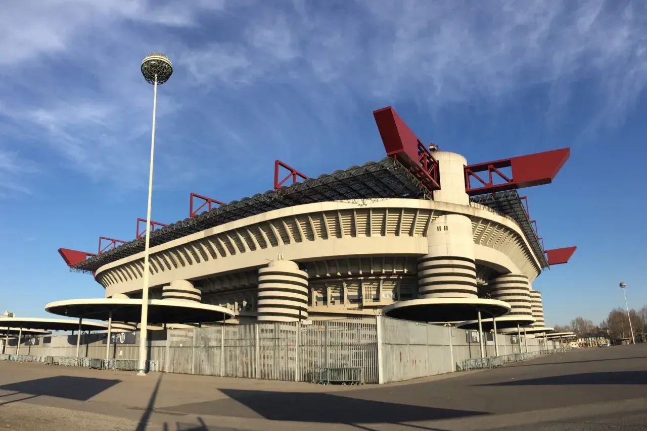 Stadion AC Milan, San Siro, z charakterystycznymi czerwonymi elementami dachu i kulistą latarnią, prezentuje się imponująco na tle błękitnego nieba.