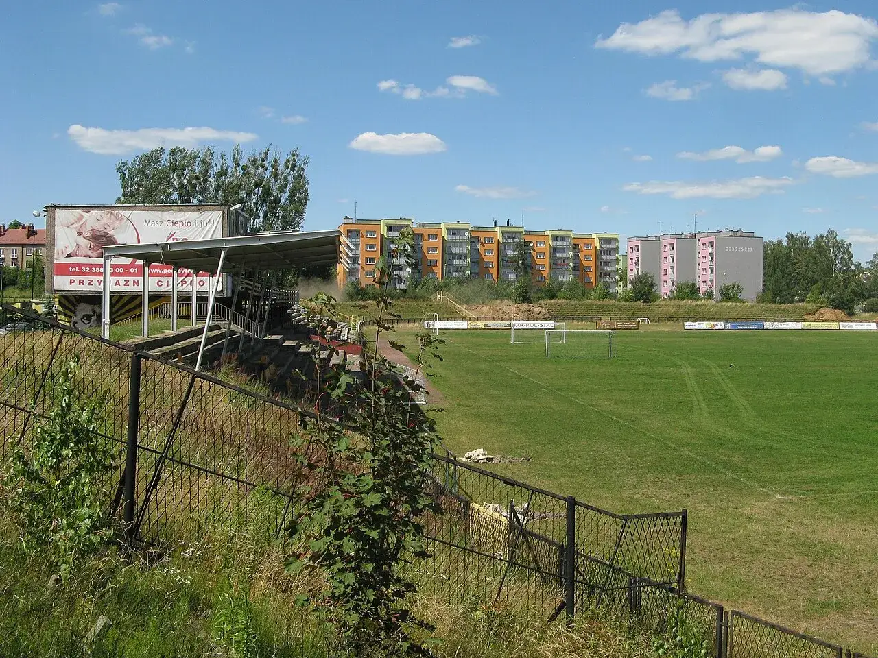 Stadion Ruchu Radzionków z trybunami i boiskiem piłkarskim. W tle bloki mieszkalne.
