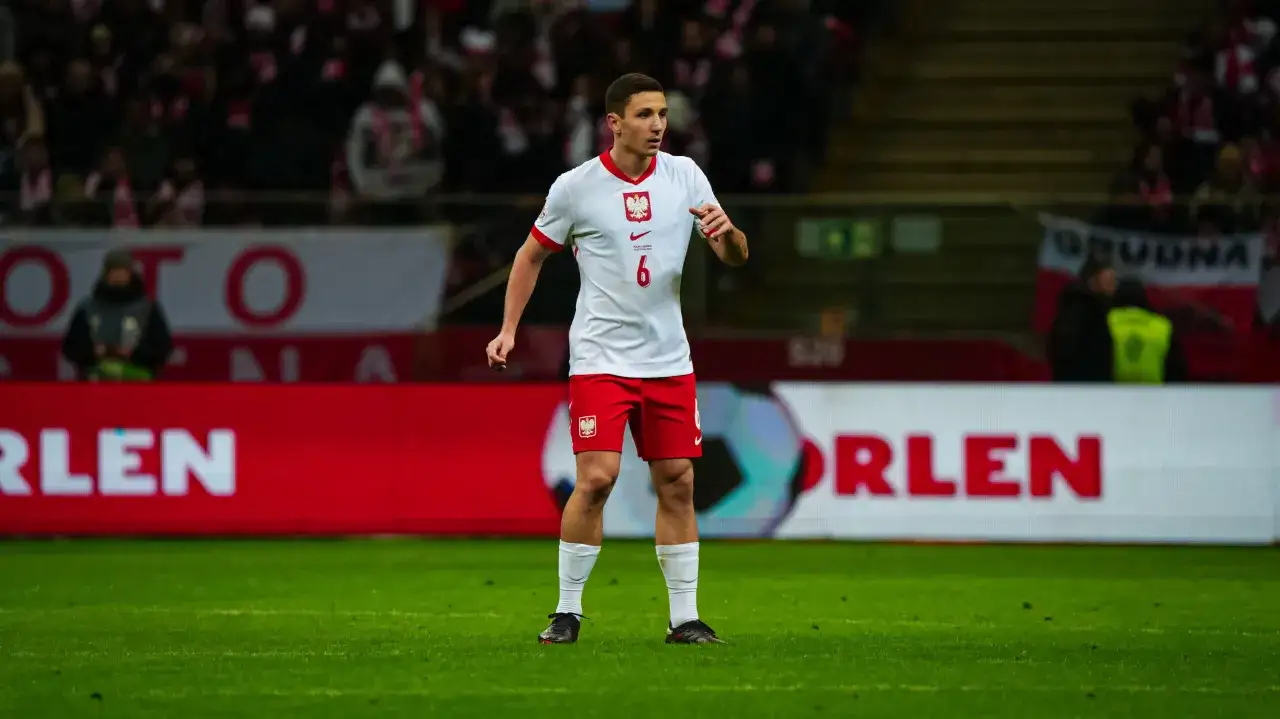 Bartosz Slisz w białej koszulce reprezentacji Polski z numerem 6, gotowy do gry na zielonej murawie stadionu.