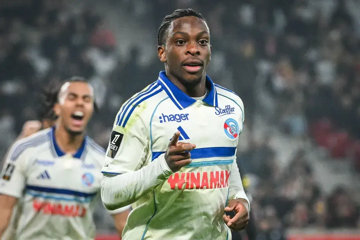 Piłkarz RC Strasbourg w białej koszulce z niebieskimi pasami, radośnie wskazuje palcem. W tle kolega z drużyny świętuje.