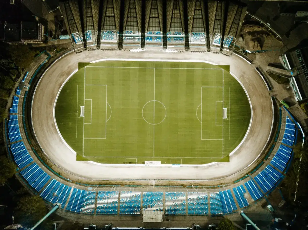 Widok z lotu ptaka na stadion miejski w Rzeszowie. Zielona murawa z białymi liniami boiska, otoczona bieżnią i trybunami z niebieskimi siedzeniami.