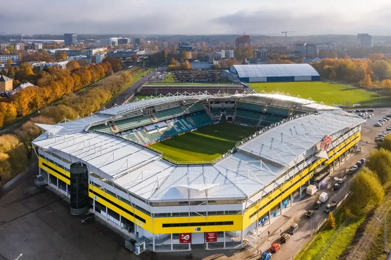 Widok z lotu ptaka na nowoczesny stadion A. Le Coq Arena z zieloną murawą i trybunami. Otoczony jesienną zielenią i zabudowaniami miejskimi.