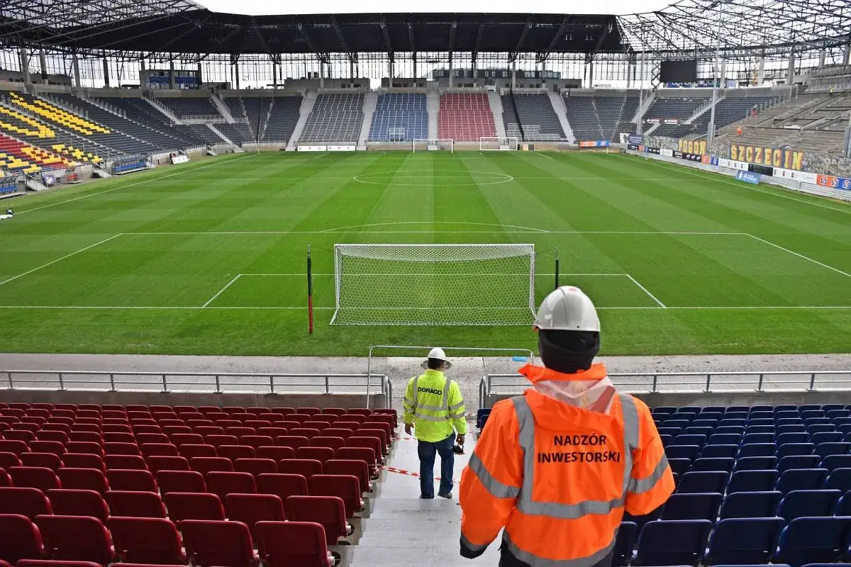 Pusta trybuna stadionu Pogoni Szczecin, z dwoma pracownikami w kamizelkach odblaskowych.