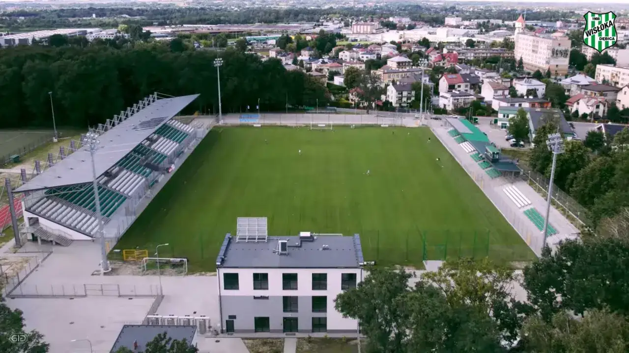 Stadion Wisłoki Dębica z zieloną murawą i trybunami, otoczony drzewami i zabudowaniami miasta.