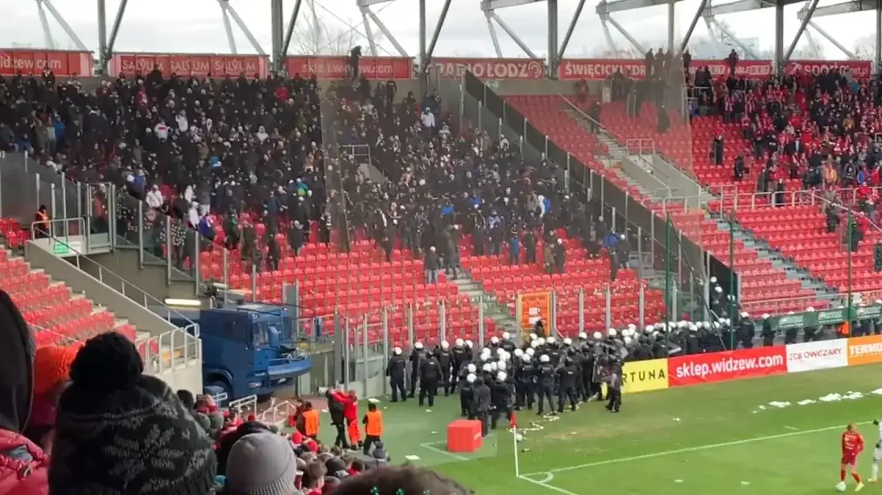 Na stadionie GKS Tychy policja interweniuje wśród kibiców. Tłum na trybunach, czerwone krzesełka, niebieski wóz policyjny.