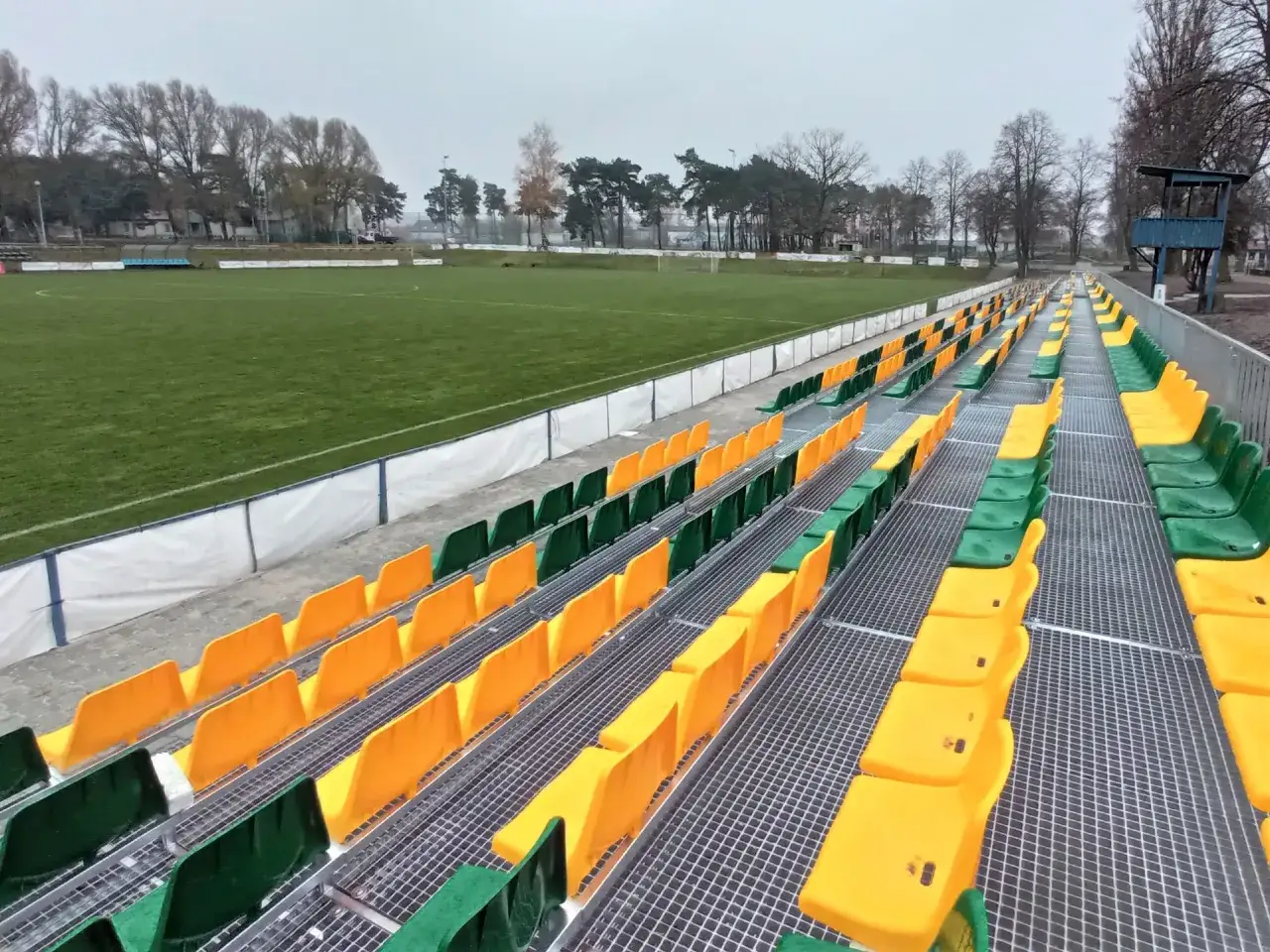 Puste trybuny stadionu Lechii Zielona Góra, z rzędami zielonych i żółtych krzesełek, czekają na kibiców.