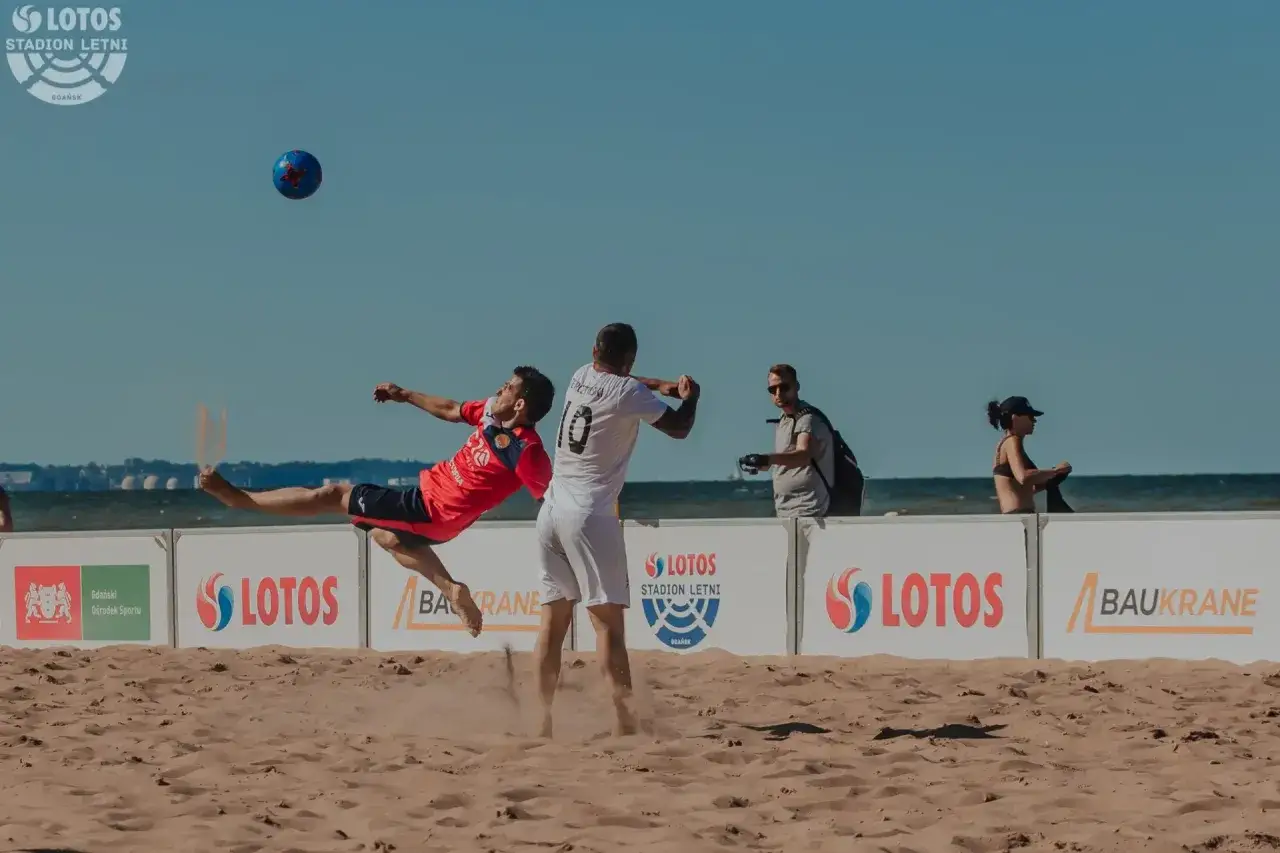 Piłkarz w czerwonej koszulce wykonuje przewrotkę na plaży podczas meczu, który może być częścią mistrzostw świata 2024.