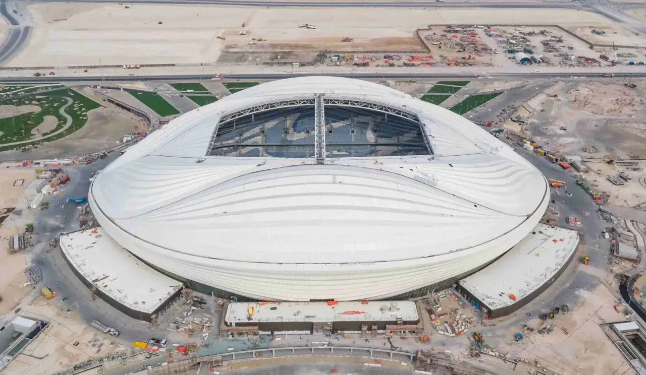 Widok z lotu ptaka na Al Janoub Stadium, nowoczesny stadion z falistym, białym dachem, otoczony placem budowy i terenami zielonymi.