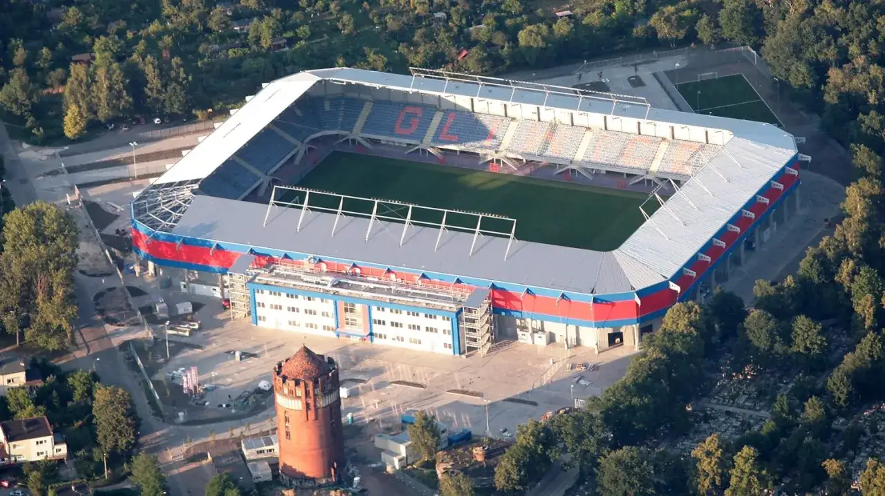 Widok z lotu ptaka na stadion Piasta Gliwice z zieloną murawą i trybunami. Obok zabytkowa wieża ciśnień.