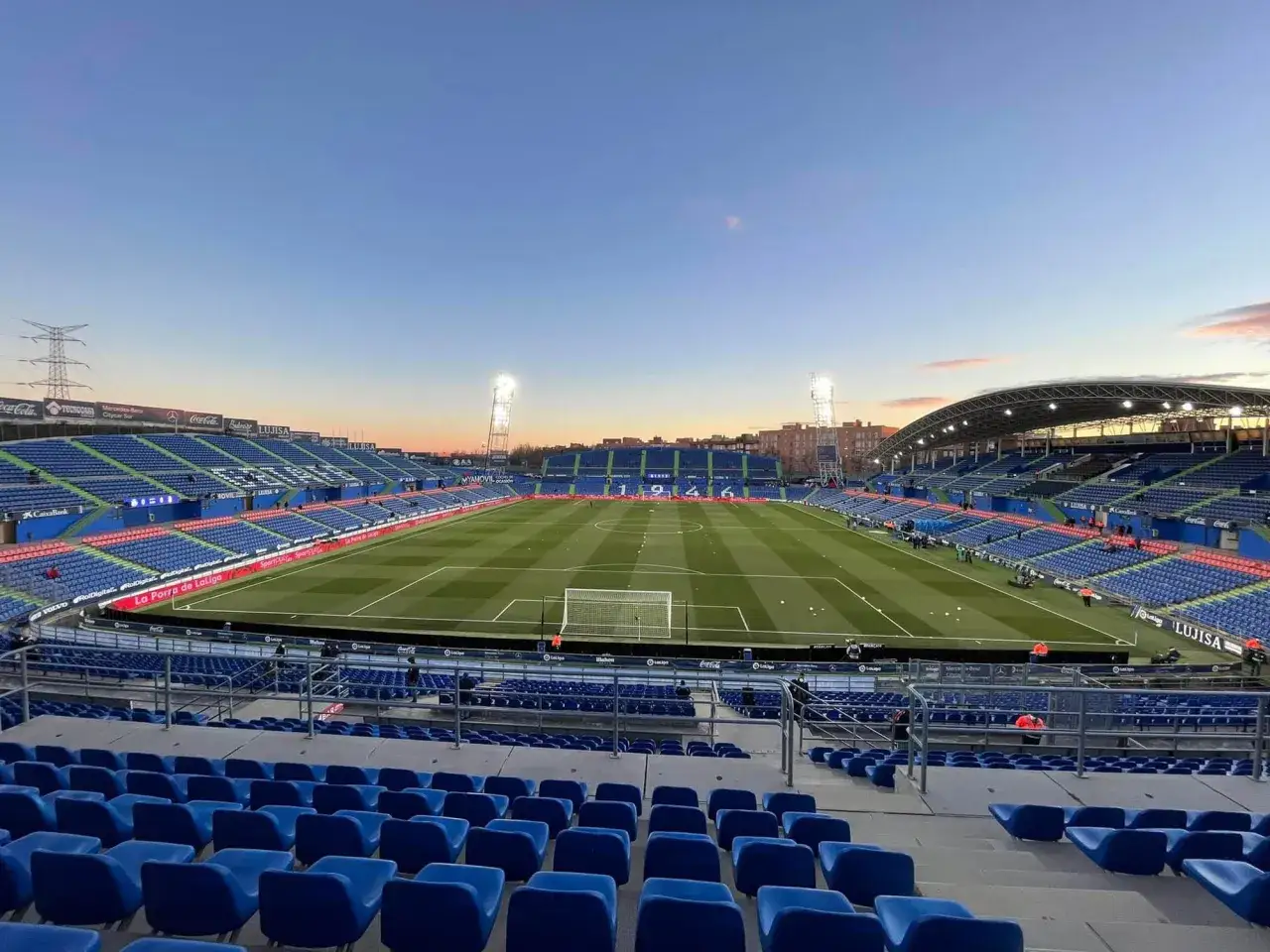 Puste trybuny stadionu Coliseum Alfonso Pérez o zmierzchu, z oświetlonym boiskiem i niebieskimi siedzeniami.