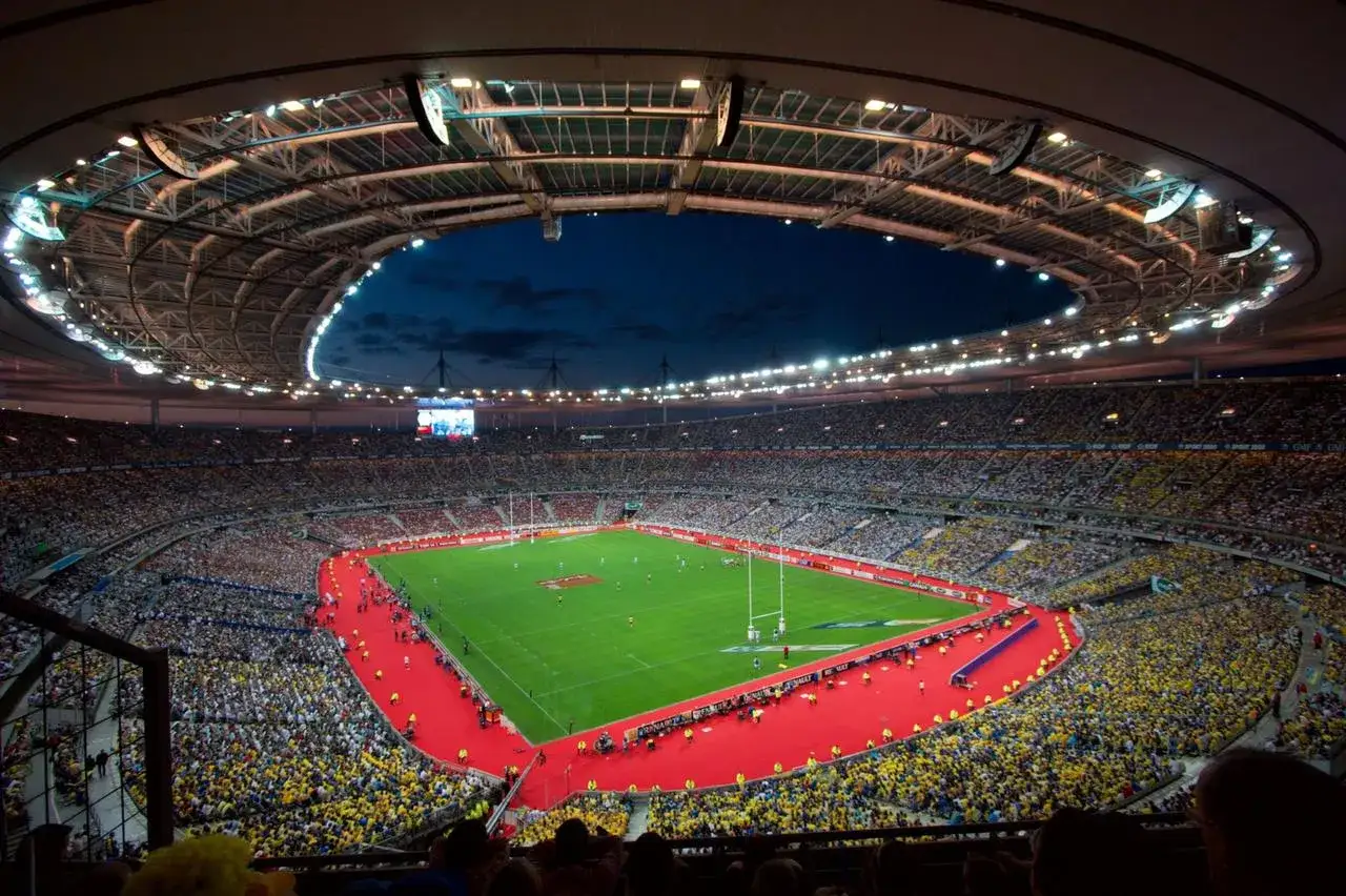 Stade de France nocą, pełne trybuny kibiców w żółtych i białych barwach, zielona murawa z czerwonym dywanem.