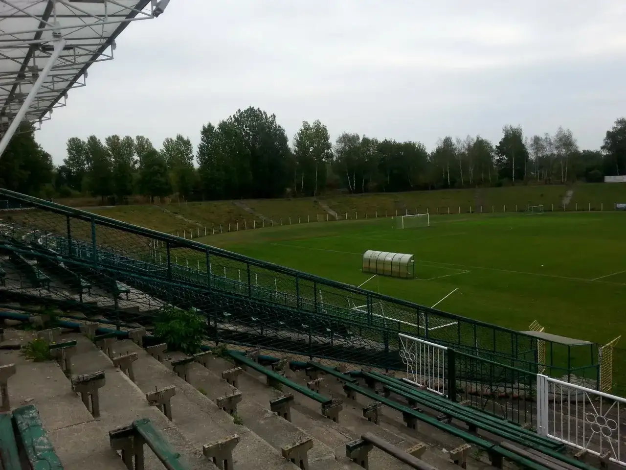 Puste trybuny stadionu Szombierki Bytom z widokiem na boisko i drzewa w tle.