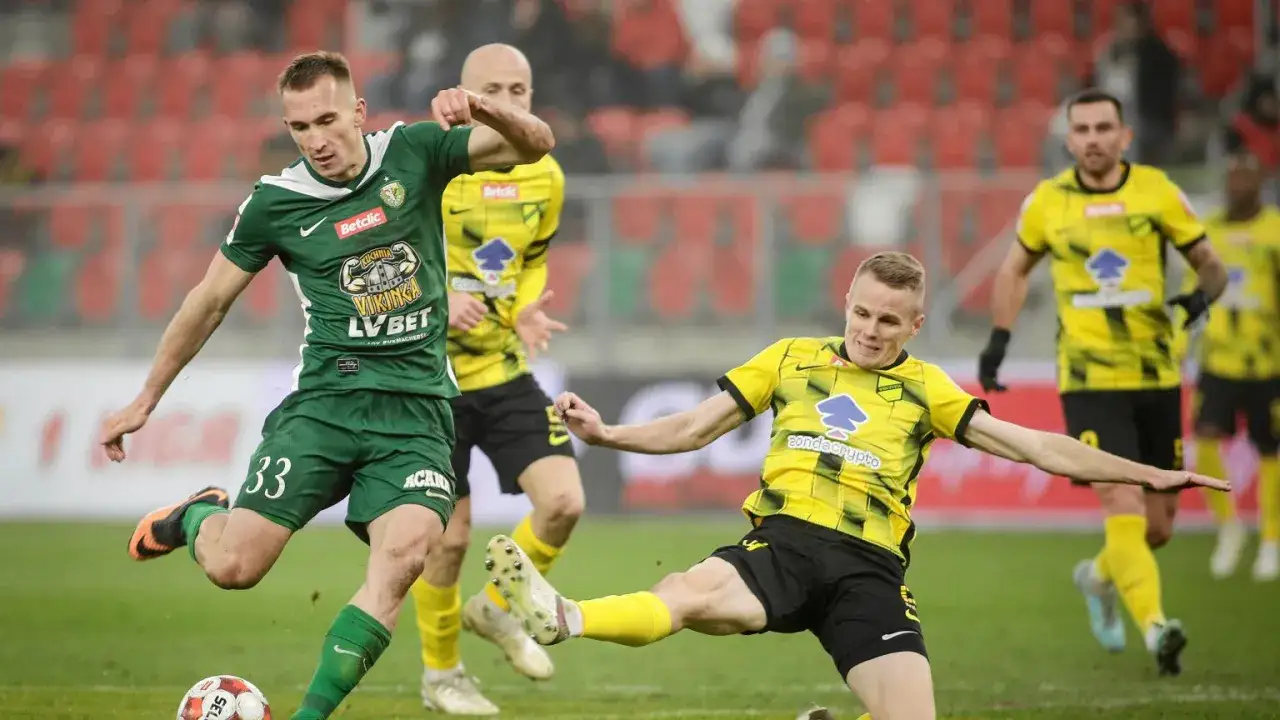 Piłkarze w akcji na boisku podczas meczu baraze 1 liga. Jeden z zawodników w zielonej koszulce kopie piłkę, a rywal w żółtej próbuje go zatrzymać.