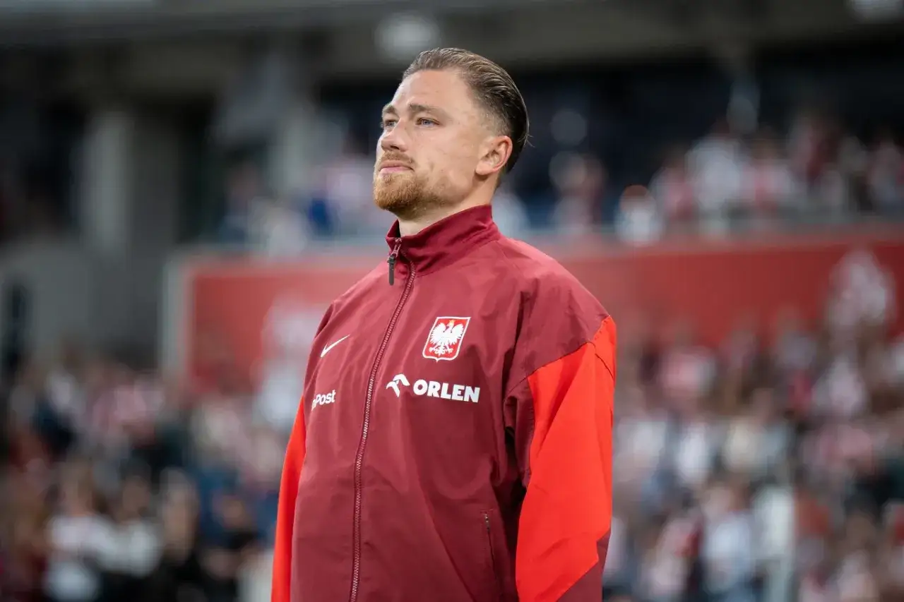 Piłkarz w bordowej kurtce z orłem na piersi, na tle trybun.