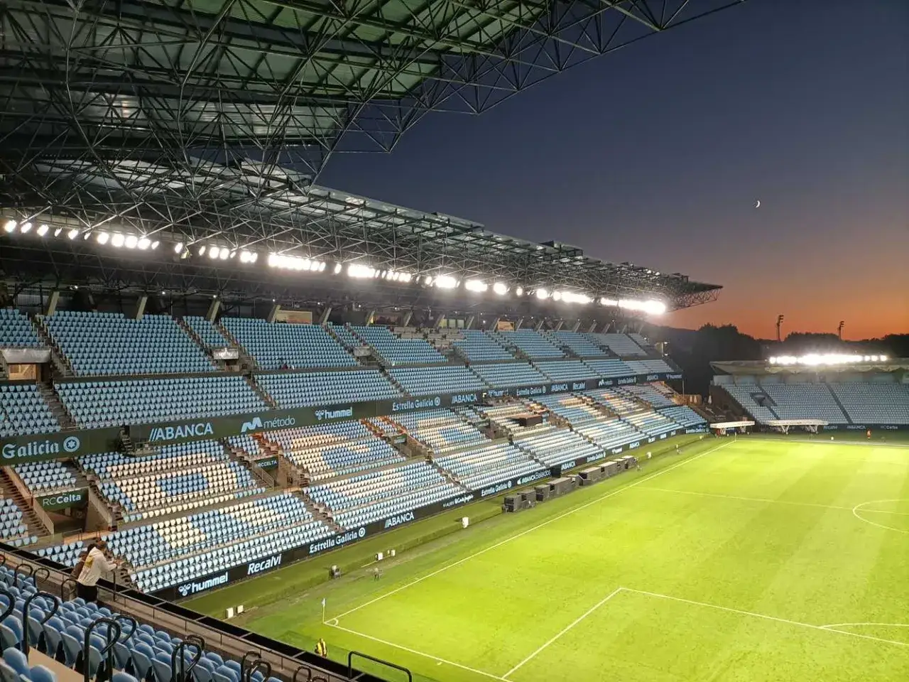 Puste trybuny stadionu Celta Vigo o zmierzchu, oświetlone jupiterami. Widać zieloną murawę i łagodne światło zachodzącego słońca.