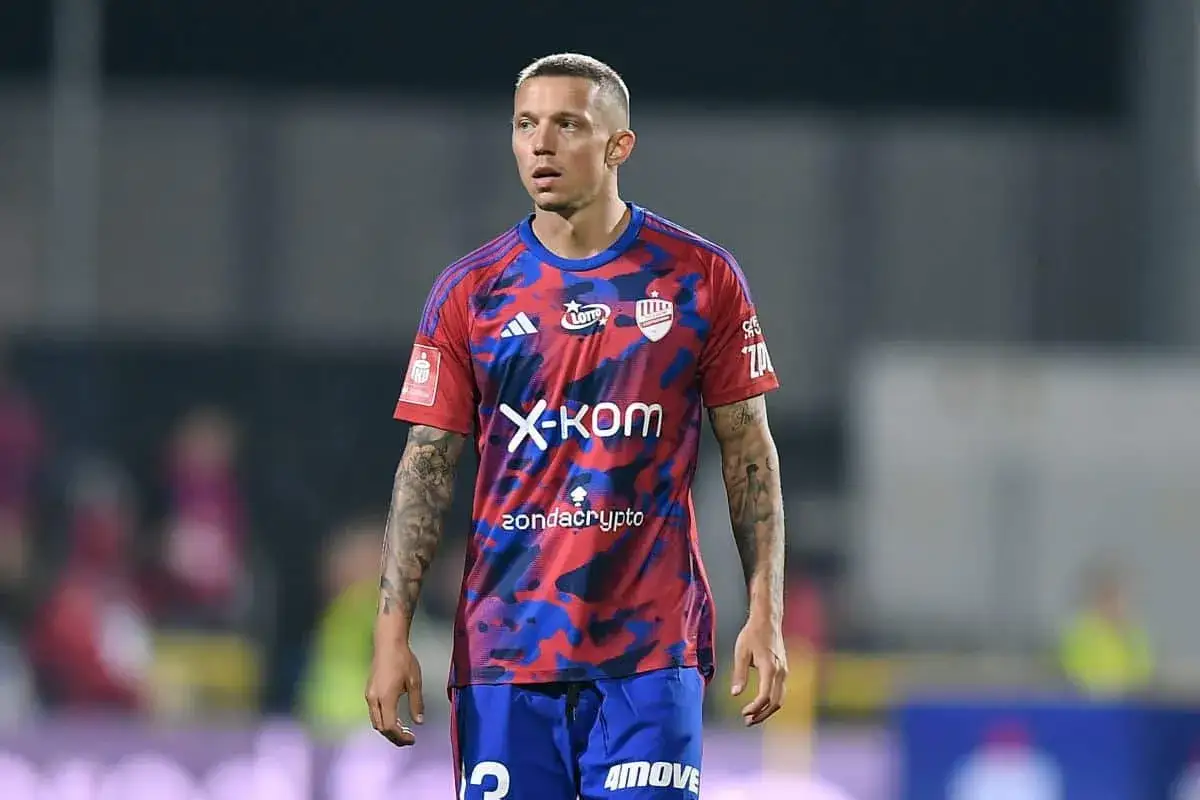 Piłkarz Sonny Kittel w czerwono-niebieskiej koszulce z logo X-KOM i tatuażami na ramionach, skupiony na boisku.