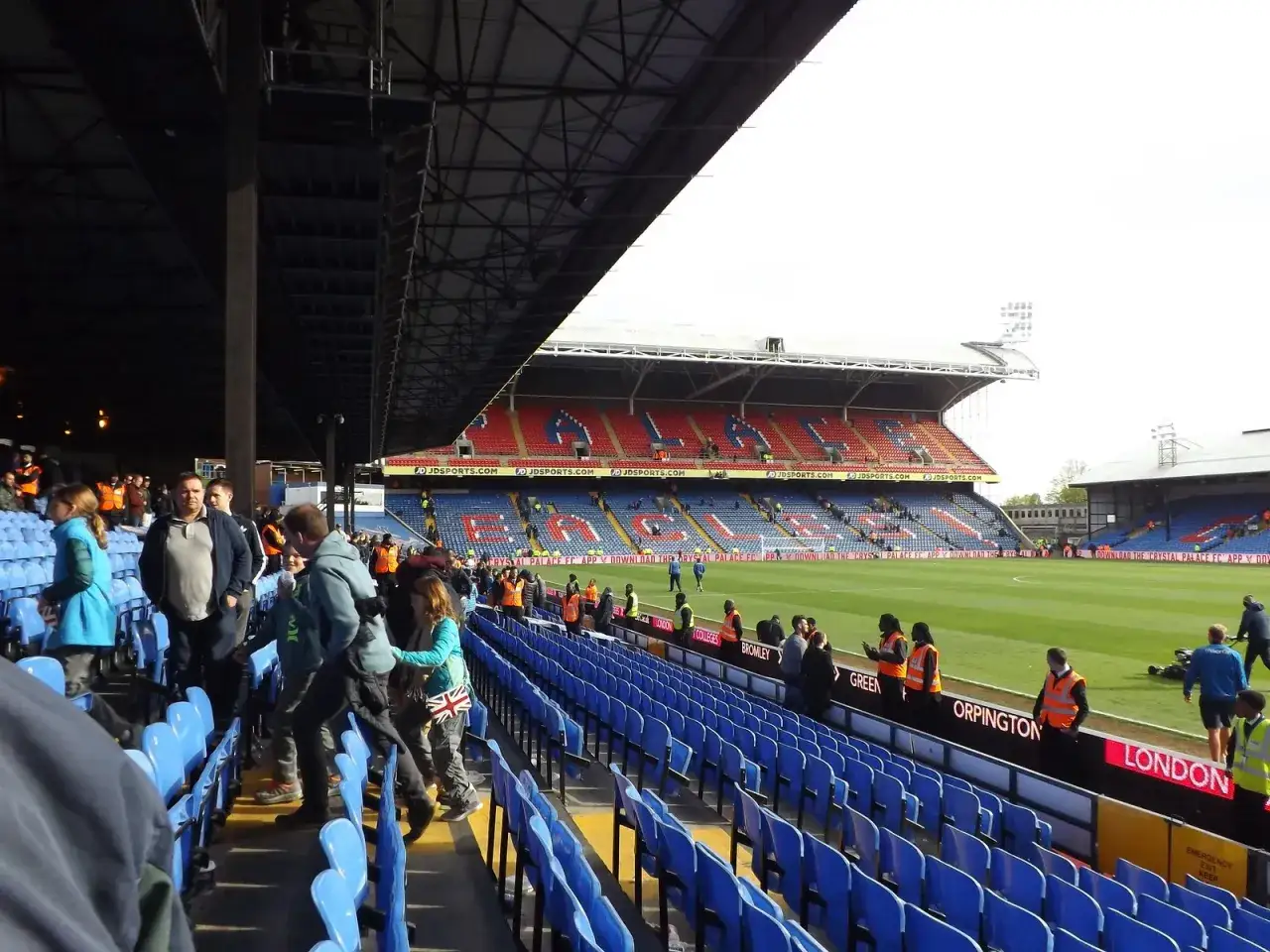 Widok na trybuny stadionu Crystal Palace z niebieskimi i czerwonymi siedzeniami. Widać fragment murawy i kibiców.