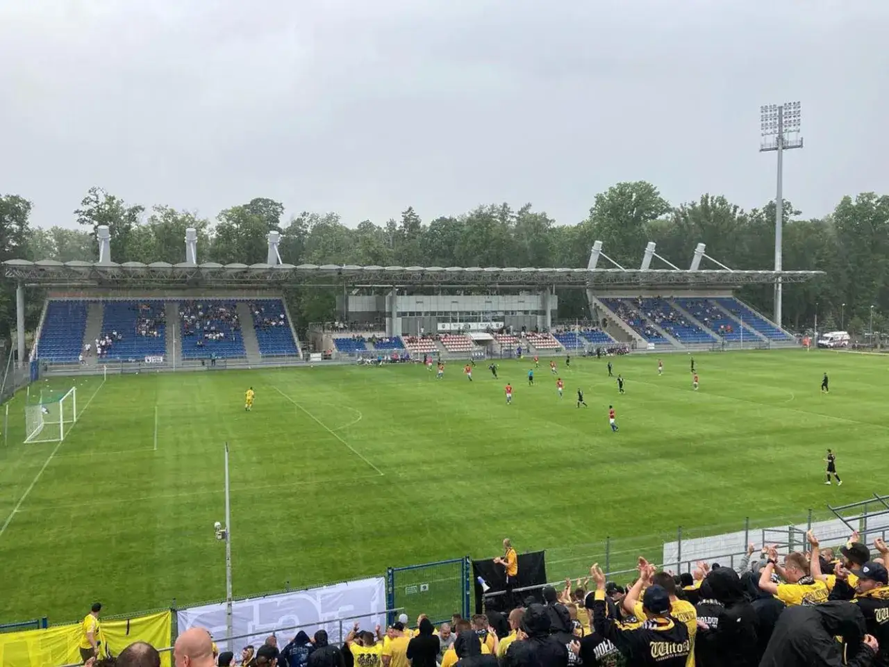 Mecz piłki nożnej na stadionie Sokoła Ostróda. Piłkarze biegają po zielonej murawie, a kibice dopingują z trybun.