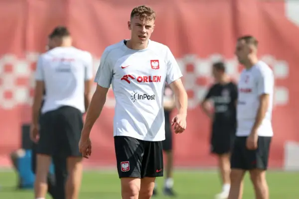 Szymon Żurkowski w białej koszulce reprezentacji Polski z logo Orlen i InPost, na tle czerwonej bandy.